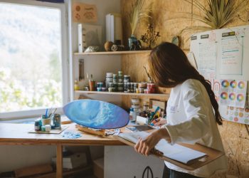 Woman artist painting a blue ocean scene on a skateboard in a cozy art studio.