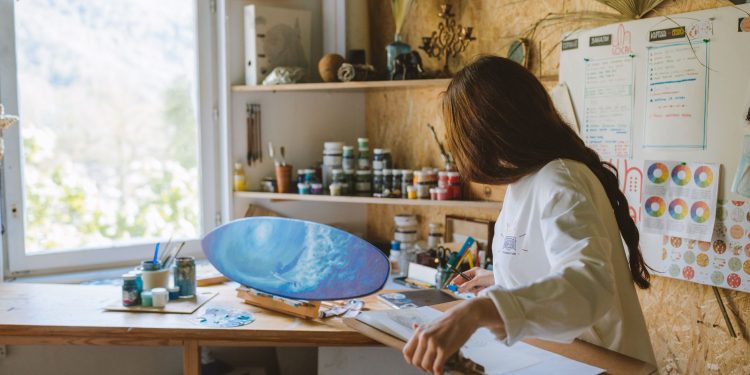 Woman artist painting a blue ocean scene on a skateboard in a cozy art studio.