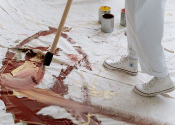 An artist uses a brush to paint abstract patterns on a large canvas placed on the floor.