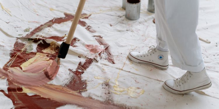 An artist uses a brush to paint abstract patterns on a large canvas placed on the floor.