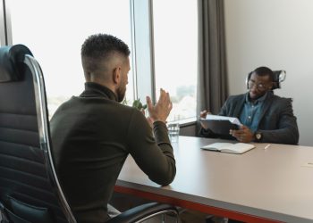 Two men in a job interview setting in a modern office with large windows.