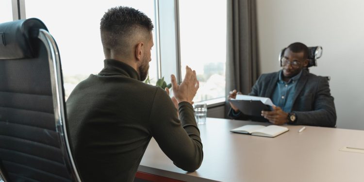 Two men in a job interview setting in a modern office with large windows.