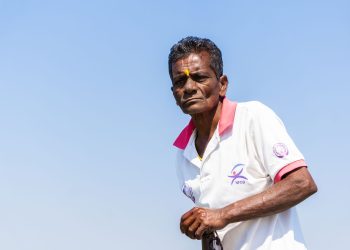 Elderly man wearing a white polo shirt with a tika stands against a clear blue sky in Chilika, India.
