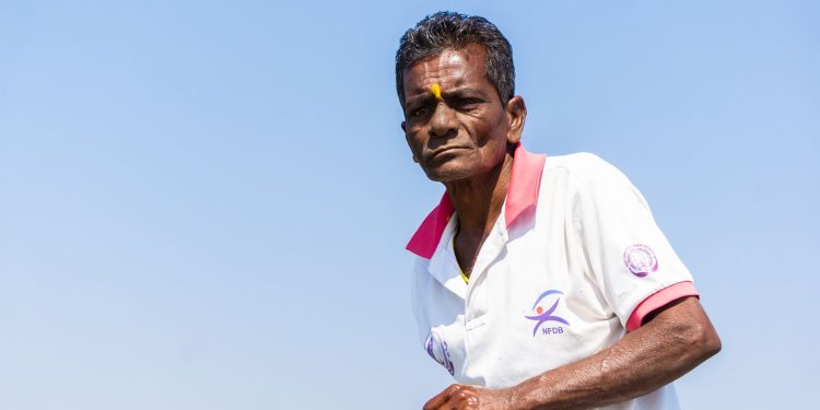 Elderly man wearing a white polo shirt with a tika stands against a clear blue sky in Chilika, India.