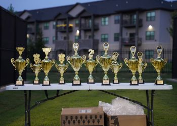 Lineup of gold trophies on a table outdoors with apartment backdrop.
