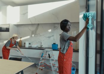 Two women cleaning a modern kitchen, wiping glass surfaces and mopping the floor.