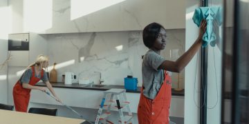 Two women cleaning a modern kitchen, wiping glass surfaces and mopping the floor.