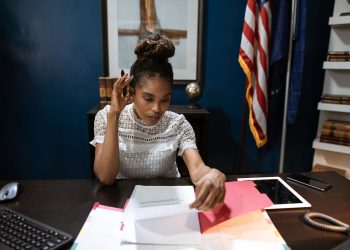 Businesswoman examines reports at desk with American flag in background.