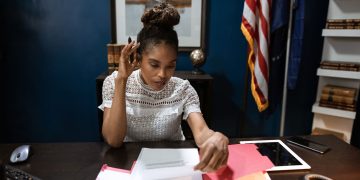 Businesswoman examines reports at desk with American flag in background.