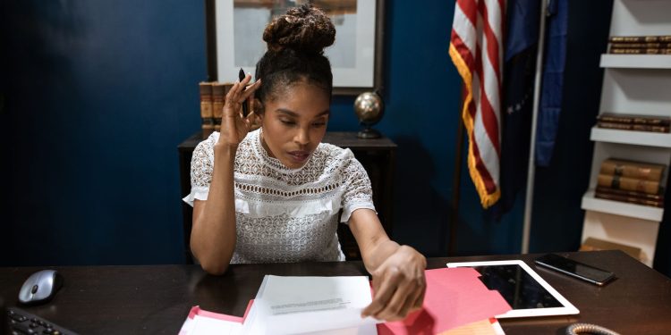 Businesswoman examines reports at desk with American flag in background.