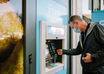 A man wearing a mask using an ATM machine outdoors for cash withdrawal.