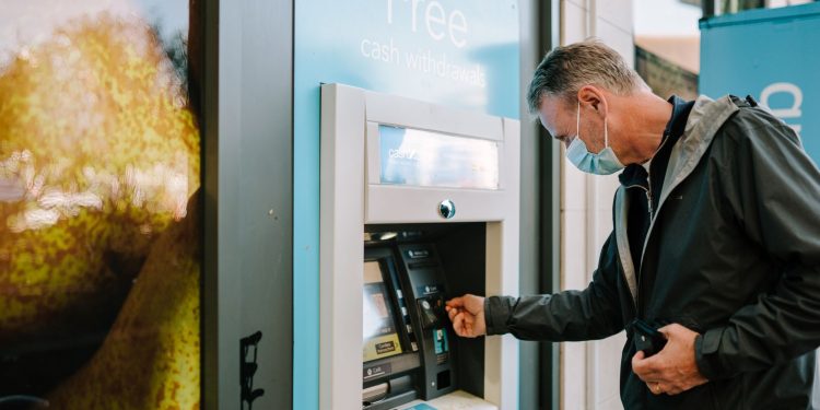 A man wearing a mask using an ATM machine outdoors for cash withdrawal.