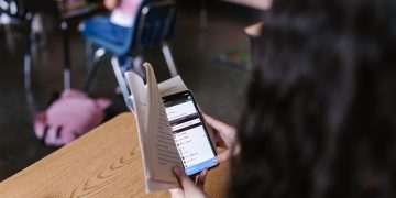 A student in a classroom multitasks by reading a book while using a smartphone, emphasizing modern educational challenges.