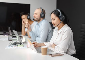 Call center agents wearing headsets working on laptops at a customer support team meeting.
