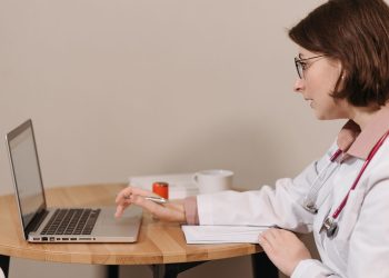 A female doctor in a white coat using a laptop while writing notes at a wooden table.