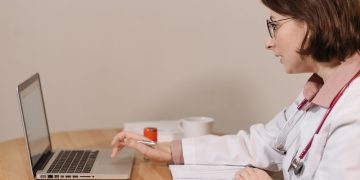 A female doctor in a white coat using a laptop while writing notes at a wooden table.