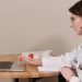 A female doctor in a white coat using a laptop while writing notes at a wooden table.