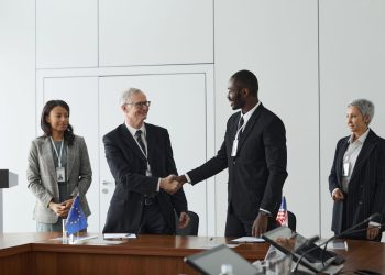 Business professionals shaking hands in conference room with flags signifying international agreement.