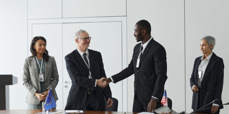 Business professionals shaking hands in conference room with flags signifying international agreement.