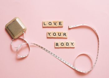 Scrabble letters spelling 'Love Your Body' with a tape measure on a pink backdrop.