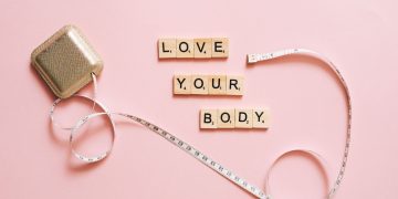 Scrabble letters spelling 'Love Your Body' with a tape measure on a pink backdrop.