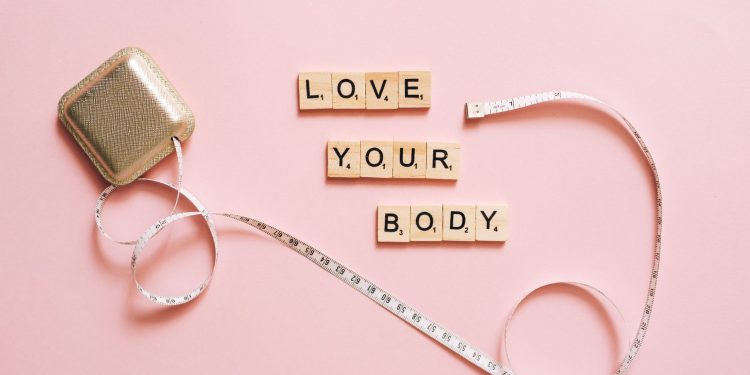 Scrabble letters spelling 'Love Your Body' with a tape measure on a pink backdrop.