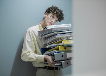 Young man overwhelmed with a stack of folders symbolizes workplace stress.