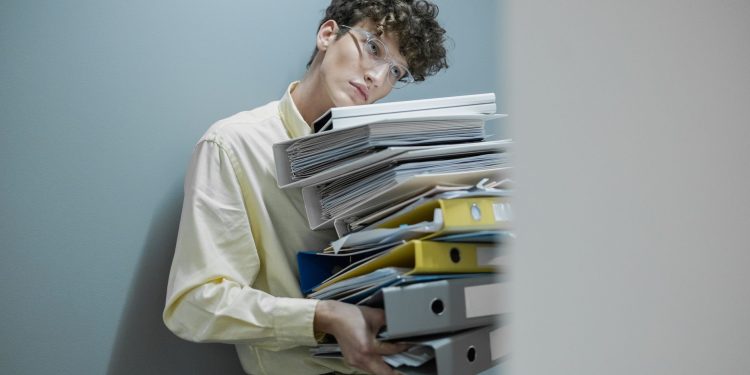 Young man overwhelmed with a stack of folders symbolizes workplace stress.