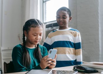 Happy little African American boy standing near Asian classmate girl using smartphone sitting at desk during break