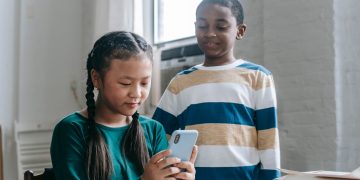 Happy little African American boy standing near Asian classmate girl using smartphone sitting at desk during break