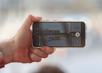 A hand holding a smartphone recording a video of a car scene outdoors. Captured in Balatonfőkajár, Hungary.