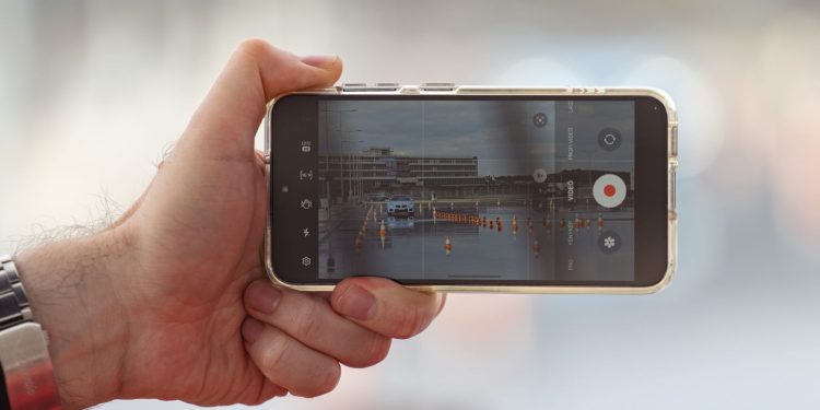 A hand holding a smartphone recording a video of a car scene outdoors. Captured in Balatonfőkajár, Hungary.
