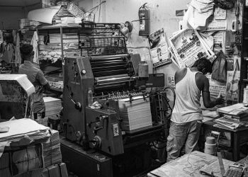 Black and white image of a bustling print shop with workers operating a printing press.