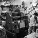 Black and white image of a bustling print shop with workers operating a printing press.