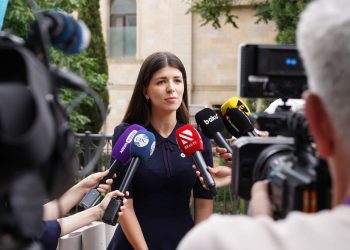 A spokesperson engages with media during an outdoor press interview surrounded by microphones.