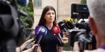 A spokesperson engages with media during an outdoor press interview surrounded by microphones.