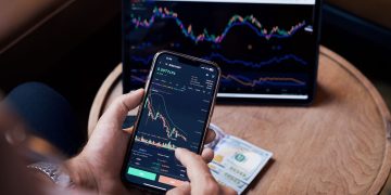 Close-up of a person trading stocks using a smartphone and a tablet.