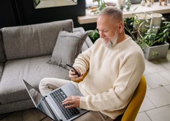A smiling elderly man sits comfortably indoors, using both a phone and laptop, with a cozy ambiance.