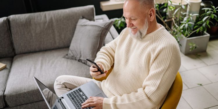 A smiling elderly man sits comfortably indoors, using both a phone and laptop, with a cozy ambiance.