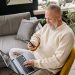 A smiling elderly man sits comfortably indoors, using both a phone and laptop, with a cozy ambiance.