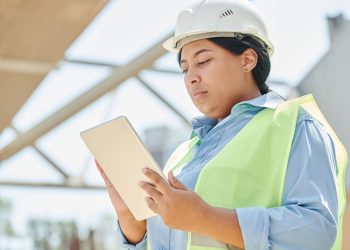 Confident female engineer in hard hat using a tablet computer at an outdoor construction site.