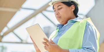 Confident female engineer in hard hat using a tablet computer at an outdoor construction site.