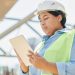Confident female engineer in hard hat using a tablet computer at an outdoor construction site.