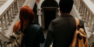 Two university students with backpacks walk down a grand staircase in a historic building.