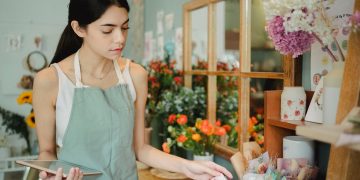 Concentrated female florist with dark hair standing in floral shop with tablet near flowers in wrapper while checking order during work