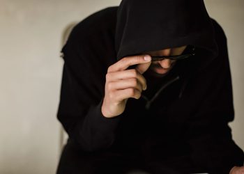 Crop focused male hacker covering head with hood while browsing laptop and lowering eyeglasses in surprise