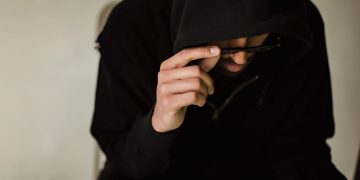 Crop focused male hacker covering head with hood while browsing laptop and lowering eyeglasses in surprise