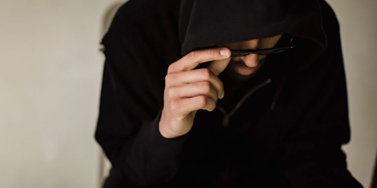 Crop focused male hacker covering head with hood while browsing laptop and lowering eyeglasses in surprise