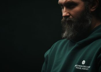 Thoughtful bearded man wearing a dark green hoodie in a moody, low-light setting.