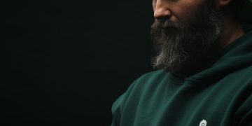 Thoughtful bearded man wearing a dark green hoodie in a moody, low-light setting.